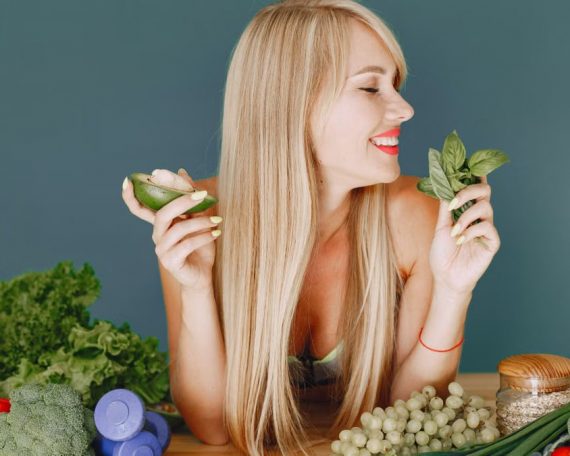 A smiling woman holding avocado and basil, surrounded by fresh vegetables and fruits for healthy eating.